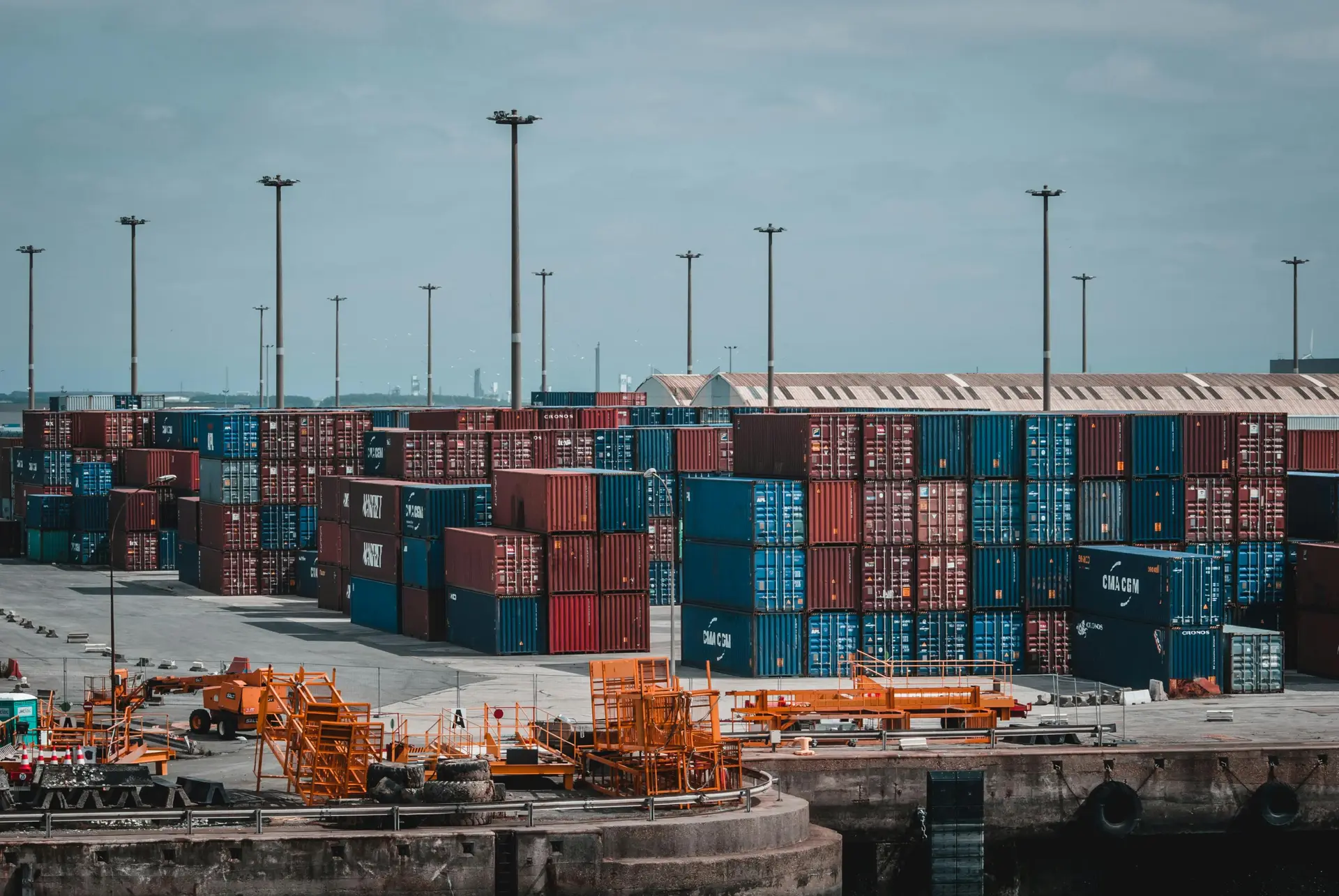 Cinematic wide shot of a shipping container terminal at blue hour with cool atmospheric lighting and glowing orange industrial safety lights